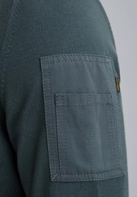 Dunkelblauer Pullover mit einem strukturierten Strickkörper und einer soliden Stoffeinfassungs-Taschen auf dem oberen Ärmel. Genähte Details sind an den Taschenkanten sichtbar.