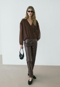 Junge Frau mit dunkler Sonnenbrille, durchsichtiger brauner Bluse, brauner Lederhose, schwarzen Ballerinas und einer kleinen schwarzen Handtasche vor neutralem Hintergrund.