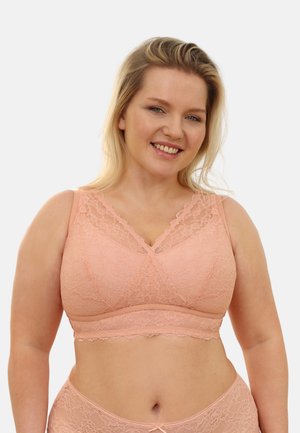 Smiling woman wearing peach lace bralette and matching underwear, standing against a plain light background.