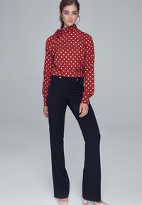 Kobieta stojąca w czerwonej bluzce w kropki z kokardą u szyi i czarnych spodni wysokich w talii na jednolitym szarym tle.