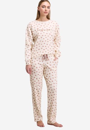 Kvinde iført cremefarvet ribbet pyjamassæt med små brune hjerter og teksten "happiness" på toppen, står barfodet med hænderne i lommerne.