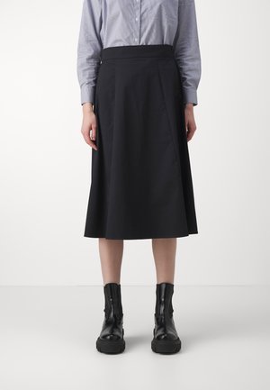 Henkilö, jolla on musta midi-hame, vaaleansininen nappipaita ja mustat nahkaiset nilkkurit, seisoo yksinkertaisen vaalean taustan edessä.