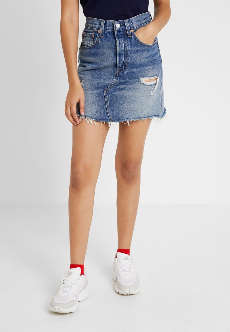 Femme portant une jupe en jean bleue usée avec un ourlet effiloché, des chaussettes rouges aux chevilles, des baskets blanches et un haut noir, debout, les mains dans les poches.