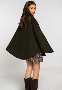 Cape en laine verte au design fluide, avec des bords arrondis et un col montant, assortie à une jupe à motifs et des bottes en daim marron.