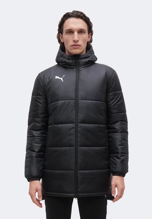 Personne portant un manteau doudoune noir à capuche avec un logo Puma blanc sur la poitrine, debout face à un fond uni.