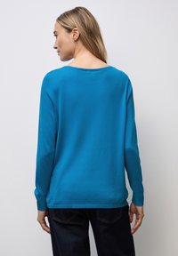 Langärmliger Pullover in leuchtendem Teal, aus weichem Stoff gefertigt. Mit Rundhalsausschnitt und gerippten Bündchen. Getragen mit dunklen Jeans.