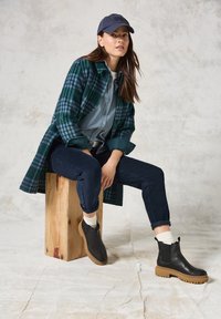 Zwarte leren enkellaarzen met een chunky tan zool, groene geruite buitenjas, lichtblauw shirt, marineblauwe broek en witte sokken. Zittend op een houten blok.