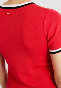 Haut rouge en maille avec manches courtes, doté d'un col rayé noir et blanc et d'un petit logo métallique à l'arrière.