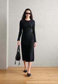 Fitted black midi dress with long sleeves, made of smooth fabric. Model carries a black bag and wears flat black shoes. Solid color design.
