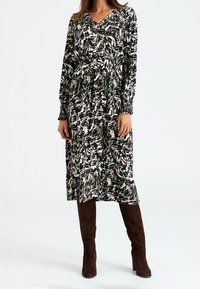 Robe à manches longues dans un motif abstrait mélangé de vert, noir et blanc. Encolure en V, poignets smockés et taille froncée. Portée avec des bottes brunes à hauteur de genoux.