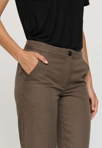 Pantalons à carreaux bruns avec une coupe ajustée, dotés d'une fermeture à bouton et de poches latérales. La texture du tissu semble lisse et soignée.