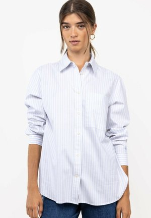 Joven mujer con cabello castaño recogido en una cola de caballo que lleva una camisa blanca con botones y finas rayas verticales y vaqueros azules.