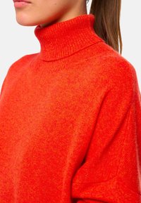 Pull à col roulé rouge en tissu doux ; il présente un col ajusté et des manches amples avec une finition texturée. Design simple et chaleureux.