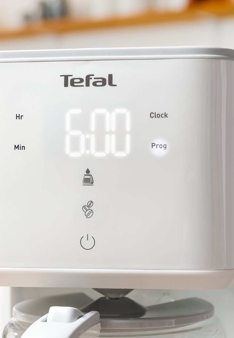Machine à café blanche avec affichage numérique indiquant "6:00". Comprend des boutons pour l'heure, la minute, l'horloge, le programme, ainsi que des icônes pour l'eau et le café.
