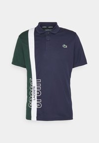 Marinblå och mörkgrön polotröja med vit vertikal rand, Lacoste-text på randen och grön krokodil-logotyp på bröstet.