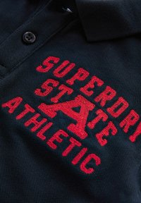 Juoda polo marškinėliai su trumpomis rankovėmis, su raudonu išsiuvinėtu tekstu "SUPERDRY STATE ATHLETIC" ir mygtuku ant apykaklės. Textūruota medžiaga.