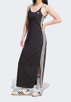 Femme portant une robe maxi noire sans manches Adidas avec des bandes blanches et un logo, des baskets blanches Adidas et des chaussettes blanches avec des bandes noires.