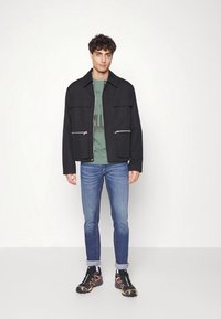 Sort lynlåsejakke med to frontlommer, båret over en grøn t-shirt, parret med blå jeans og sorte sneakers med orange detaljer.