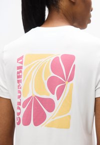 Άτομο που φοράει λευκό πουκάμισο με ροζ και κίτρινο αφηρημένο λουλουδάτο σχέδιο και τη λέξη "COLOMBIA" τυπωμένη στην πλάτη.