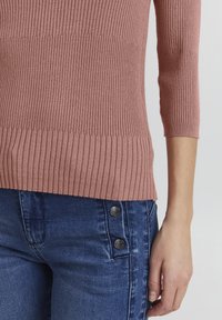 Różowy sweter w prążki z rękawami 3/4, o dopasowanym kroju. Noszony z niebieskimi dżinsami, podkreślonymi dwoma metalowymi guzikami z boku.