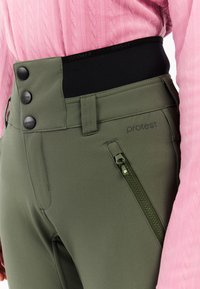 Close-up van olijfgroene high-waist broek met zwarte elastische tailleband, drie knoopsluitingen, ritszak, gedragen met een roze textuurtrui.
