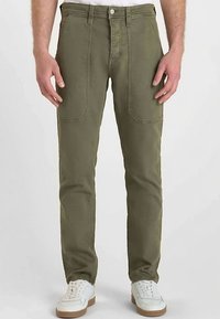 Pantalon en coton vert olive à coupe droite, avec deux poches avant et une braguette zippée, associé à des baskets blanches.