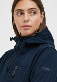 Marineblått softshell-jakke med hette, glidelåslommer og justerbare snorene. Har "NORTH BEND"-logoen på ermet.