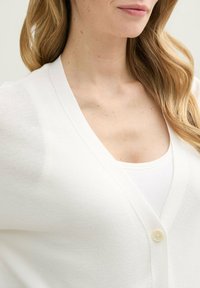 Witte cardigan met een V-hals ontwerp, lichte gebreide textuur en een enkele ronde knoopsluiting. Daaronder bevindt zich een witte tanktop.