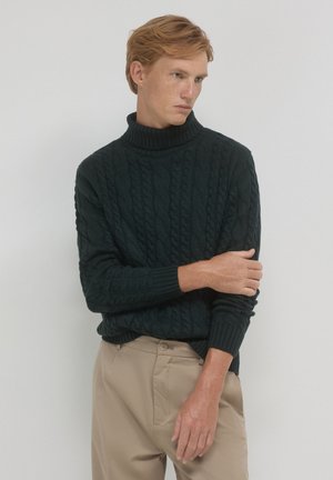 Dunkelgrüner Strukturstrick-Turtleneck-Pullover mit gerippten Bündchen und Saum, kombiniert mit hellbeigen Hosen. Glatte Textur, detailliertes Muster.