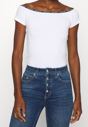 Persoon draagt een nauwsluitend, wit off-shoulder topje met het Calvin Klein Jeans-logo en high-waisted blauwe spijkerbroek met zichtbare knoopsluiting.
