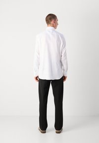 Chemise blanche à manches longues avec un col pointu, dotée d'un ourlet droit et de poignets boutonnés, assortie à un pantalon noir et des chaussures décontractées.