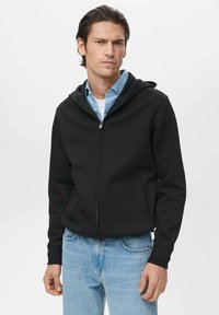 Sudadera negra con cremallera y bolsillo frontal, puños ajustados y una capucha amplia, llevada sobre una camisa de cuello azul claro y jeans de mezclilla.