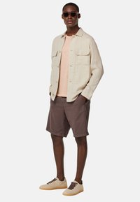 Hellbeige Leinenhemd über einem pfirsichfarbenen T-Shirt, kombiniert mit dunkelbraunen Shorts und hellbeigen Sneakers mit dunklen Schnürsenkeln. Schwarze Sonnenbrille.