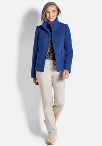 Frau, die lächelt, trägt einen blauen Knopfmantel, ein gemustertes Oberteil, beigefarbene Hosen und beigefarbene Sneaker und steht vor einem schlichten weißen Hintergrund.