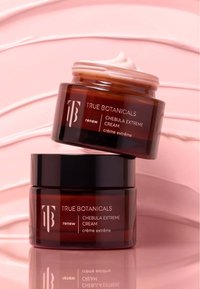 Deux pots ambrés de la Crème Extrême Chebula de True Botanicals sur un fond rose texturé, l'un ouvert avec de la crème visible.