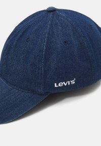 Tumši zilā džinsa cepure ar izliekto malču, priekšā ar baltu Levi's logo izšuvumu un šuvju detaļām uz auduma.
