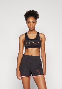 Crna sportska majica s natpisom "HYROX" u šarenim slovima. Uparena s crnim kratkim hlačama s ružičastim detaljima i logotipom na lijevoj nozi.