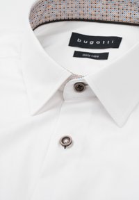 Weißes Hemd mit strukturiertem Kragen, mit gemusterter Innenseite und braunen Knöpfen. Labels zeigen Marke und Pflegehinweise an.