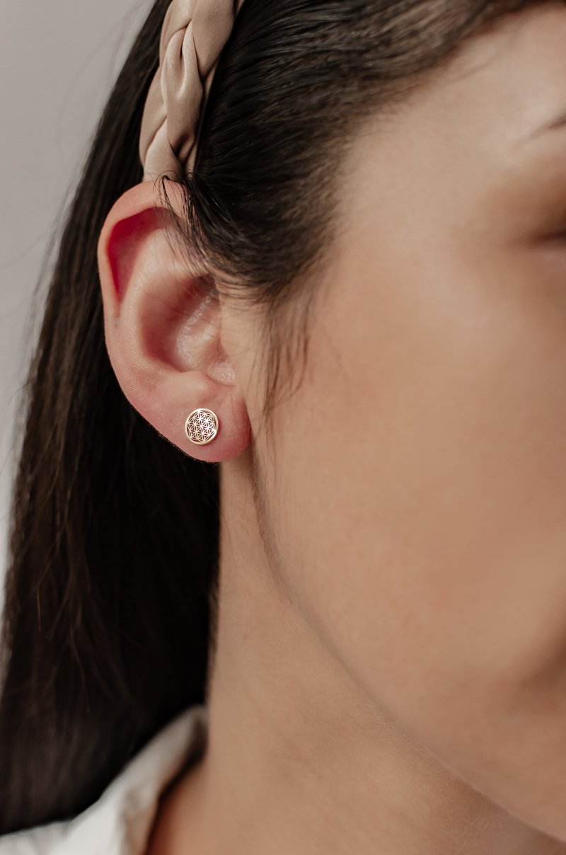 Orecchini a perno dorati con un motivo geometrico, montati su una base rotonda, indossati nel lobo dell'orecchio su pelle liscia e capelli scuri.