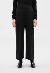 Άτομο που φοράει μαύρο ψηλόμεσο παντελόνι με φαρδύ πόδι και μαύρες μπότες με μυτερή μύτη, πάνω σε απλό λευκό φόντο.