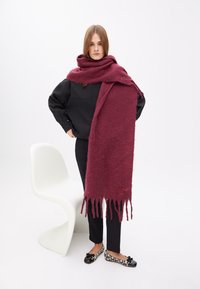 Burgundy uld tørklæde med frynser, båret over et sort outfit, med et subtilt broderet logo i hjørnet.