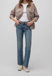 Camicia di velluto a coste color marrone chiaro con due tasche sul petto, t-shirt bianca sotto, jeans dritti blu e sneakers bianche.