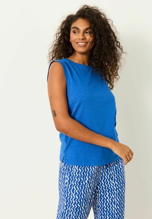 Kobieta z kręconymi włosami, ubrana w niebieski bezrękawnik i luźne spodnie w niebiesko-biały falisty wzór, uśmiechająca się i patrząca na bok.