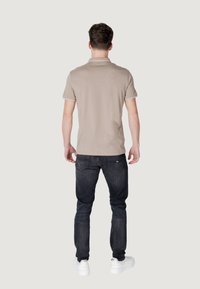 Polo beige con bordi bianchi, indossato con jeans scuri e sneakers bianche. Visto da dietro, mette in risalto il design minimalista e lo stile casual.