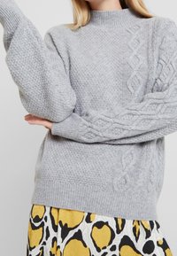 Šedý pletený svetr s vysokým límcem, zdobený copánkovými vzory na rukávech, spárovaný s sukní s žlutým, černým a bílým vzorem.