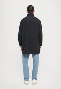 Black waterproof jacket with a high collar, relaxed fit, elastic cuffs, and side slits. Paired with light blue jeans and white sneakers.