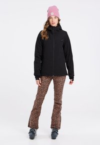 Zwarte buitenjack met ritssluiting, gecombineerd met broek met luipaardprint. Het model draagt een roze gebreide muts en zwarte skibotten.