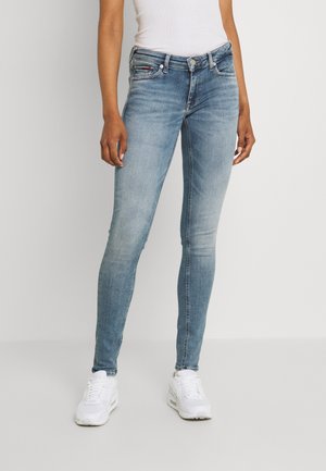Personne portant un jean slim bleu clair et des baskets blanches, debout devant un fond uni et clair.