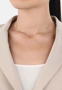 Guldhalsband med en vågig design, fin textur och delikat detaljer, bärs mot en neutralt färgad outfit. Ljus reflekteras mjukt från metallen.