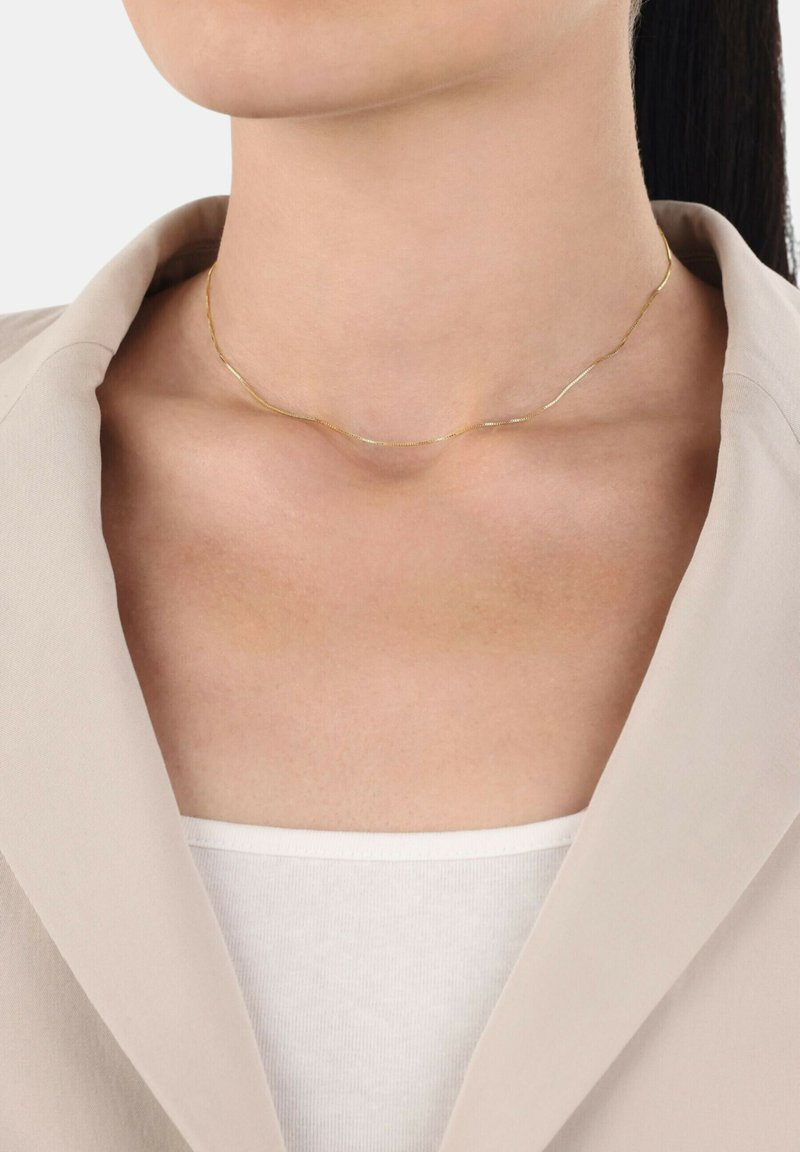 Guldhalsband med en vågig design, fin textur och delikat detaljer, bärs mot en neutralt färgad outfit. Ljus reflekteras mjukt från metallen.
